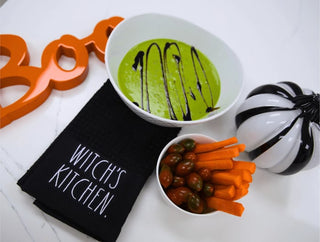 A styled Halloween-themed tabletop scene with a white bowl of bright green soup topped with dark drizzle, a small bowl of carrot sticks and olives, a black cloth napkin reading “WITCH’S KITCHEN,” an orange decorative “Boo” sign, and a black-and-white pumpkin on a white surface.