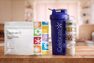 Celebrate brand supplement kit on a wooden table, featuring a white sample pouch, a dark blue shaker bottle, and a clear multi-compartment container with capsules and powder, set against a blurred kitchen background.