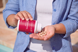 A person in a blue button-up shirt holds a white bottle with a red label, pouring beige capsules into their hand in an outdoor setting.