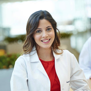A smiling woman with dark hair stands indoors wearing a white lab coat over a red top, leaning on a wooden table in a bright, softly blurred office or clinic setting.