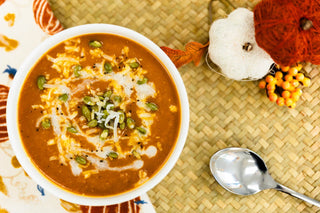 A bowl of thick orange-brown soup topped with shredded cheese, green pumpkin seeds, and a swirl of white cream, set on a woven mat with a spoon and autumn decorations nearby.