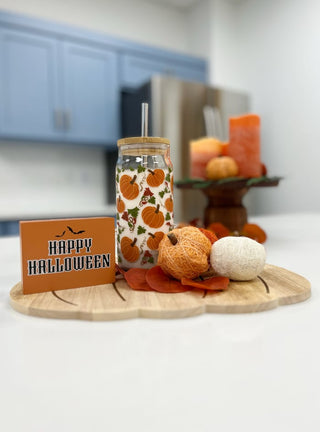 A Halloween-themed glass jar tumbler with a pumpkin pattern and a straw, styled on a wooden board with a 'Happy Halloween' sign, small pumpkins, and candles in a kitchen setting.