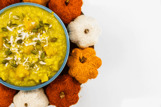 A top-down view of a blue bowl filled with yellow pumpkin spice dessert, garnished with pumpkin seeds and shredded coconut, surrounded by decorative orange and white sugar skulls on a white background.
